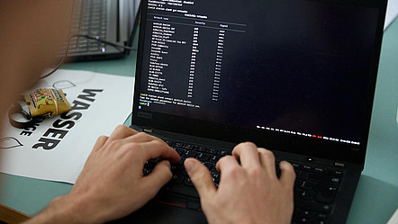 Zu sehen ist ein schwarzer Laptop mit Blick auf den Bildschirm. Im Vordergrund sind die Hände eines Menschen zu erkennen, die etwas auf der Tastatur eingeben. Der Bildschirm zeigt weiße Schrift auf einem schwarzen Hintergrund. Es wurde ein Befehl zum Anzeigen der verfügbaren WLAN-Netzwerke in der Eingabeaufforderung eingegeben.